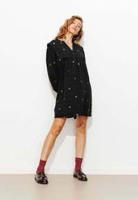 Robe noire à boutons avec manches longues et large col, ornée de motifs d'étoiles brodés. Portée avec des chaussettes bordeaux et des mocassins brillants.