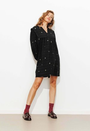 Robe noire à boutons avec manches longues et large col, ornée de motifs d'étoiles brodés. Portée avec des chaussettes bordeaux et des mocassins brillants.