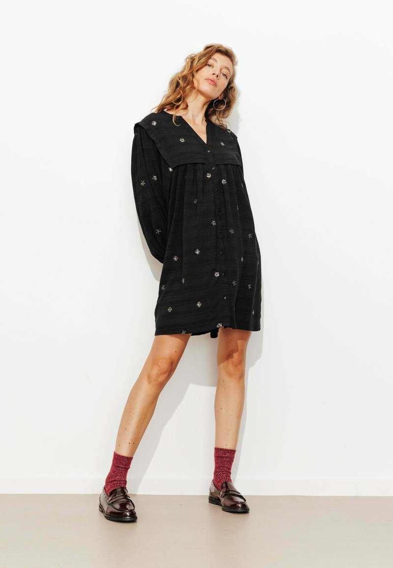 Robe noire à boutons avec manches longues et large col, ornée de motifs d'étoiles brodés. Portée avec des chaussettes bordeaux et des mocassins brillants.