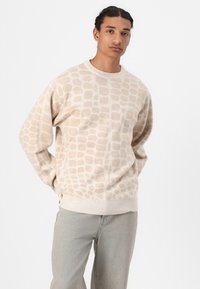 Beige gemusterter Pullover mit strukturiertem Design, gerippten Bündchen und Saum, kombiniert mit hellblauen Jeans, vor einem weißen Hintergrund.