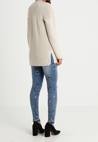 Béžový pletený svetr s žebrovanou texturou a bočními rozparky; doplněný modrými džínami s květinovou výšivkou a černými botami na blokovém podpatku.