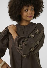 Brown button-up shirt with puffed sleeves, featuring textured floral embellishments in a matching green hue. Smooth cotton fabric.