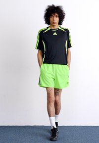 Tricou sport negru cu accente verzi neon, asortat cu pantaloni scurți verzi neon. Prezintă branding Adidas și este completat de adidași negri.