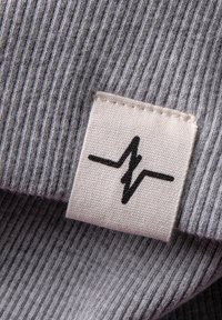 Grijs ribbelstof met zichtbare textuur. Een wit label met een zwart hartslaggrafiek is in de stof gestikt.