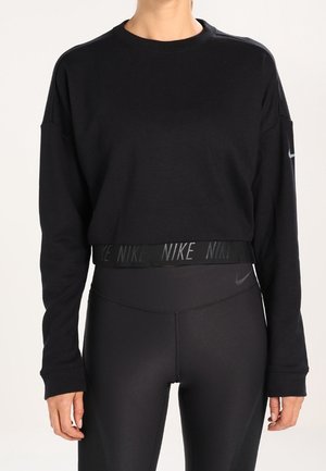 Femme portant un crop top noir à manches longues et des leggings noirs taille haute, tous deux avec des logos Nike subtils.