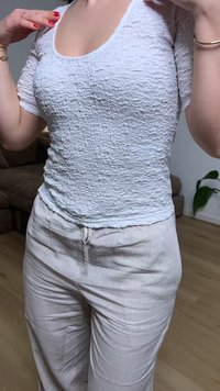 Γυναίκα ρυθμίζει λευκό κοντομάνικο τοπ με υφή, φοράει ανοιχτό μπεζ παντελόνι, με κόκκινο βερνίκι νυχιών και χρυσά βραχιόλια σε εσωτερικό χώρο.