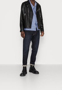 Zwarte leren motorjas, blauwe gestreepte shirt, donkere spijkerbroek en zwarte gevechtslaarzen met stevige zolen en opgerolde mouwen.