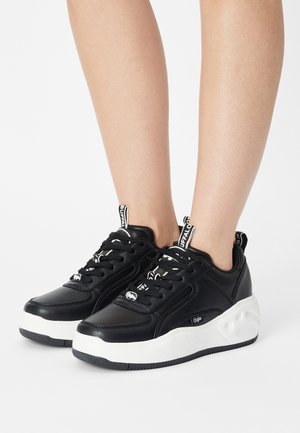 Musta nahkainen lenkkikenkä, jossa on valkoinen kuvioitu pohja, nauhallinen malli ja näkyvät logokoristeet kielen ja kannan kohdalla.