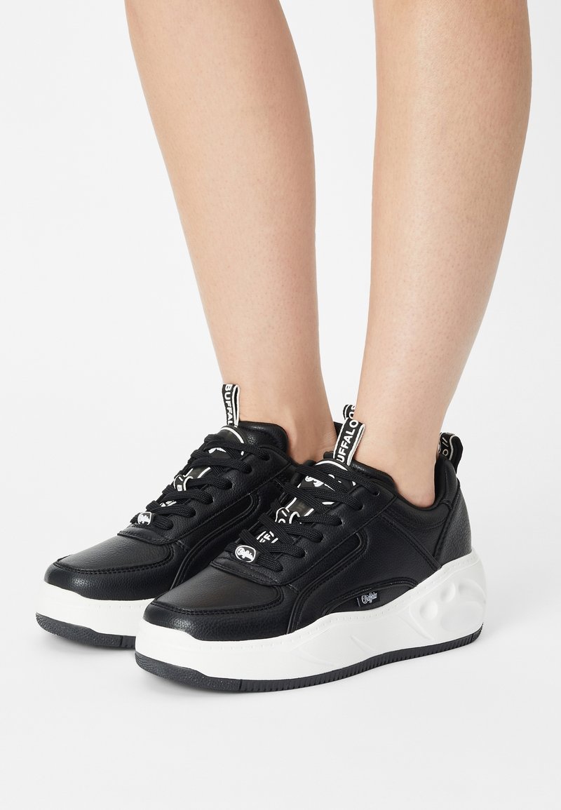 Baskets en cuir noir avec une semelle blanche texturée, design à lacets et accents de logo visibles sur la languette et le talon.