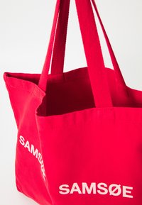Red canvas tote bag with two shoulder straps. Features white printed text reading "SAMSØE" on the front. Open top, unstructured design.