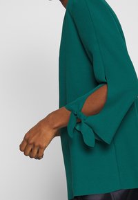 Blouse turquoise à manches trois-quarts avec des ouvertures nouées aux poignets. Fabriquée dans un tissu lisse avec une coupe ample et décontractée.
