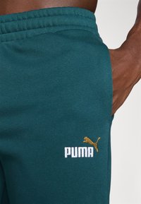 Pantalones de chándal en color teal hechos de tela suave, con un logotipo bordado en blanco de "PUMA" y un logotipo dorado de un gato. Cintura elástica sobre una textura lisa.