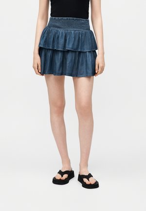 Femme portant une mini-jupe en jean bleu à volants avec une taille smockée, un top sans manches noir et des tongs plateformes noires, debout sur un fond blanc.
