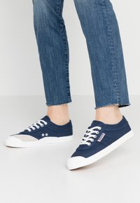 Marineblaue Canvas-Sneaker mit weißer Gummisohle, weißen Schnürsenkeln und einem hellgrauen Akzent. Verfügt über Ösen und ein strukturiertes Design im Obermaterial.