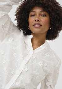 Valkoinen broderad paita med blommönster, knäppning och texturerad tyg. Designen inkluderar krage och långa ärmar.