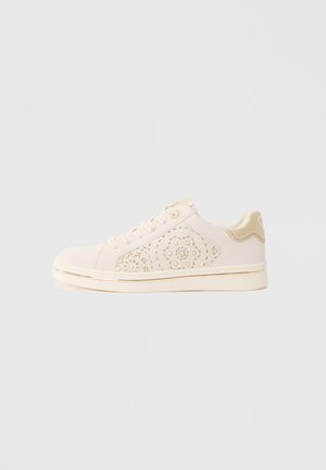 Μπεζ αθλητικό παπούτσι από καμβά με διάτρητο λουλουδάτο σχέδιο στο πλάι, χρυσές λεπτομέρειες στη φτέρνα και τη σόλα, και επίπεδο κλείσιμο με κορδόνια.