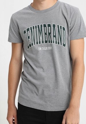 Personne portant un t-shirt gris avec les textes "DENIMBRAND" et "TOM TAILOR DENIM", debout devant un fond clair et uni.