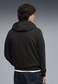 Sweat à capuche noir avec capuche doublée, poignets et ourlet côtelés, texture douce et coupe décontractée. Porté sur une chemise blanche, associé à un jean foncé.