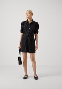Robe noire à manches courtes avec un devant à boutons et une ceinture à la taille. Le tissu a une texture lisse. Le mannequin porte un petit sac noir et des chaussures plates.
