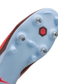 Sole di scarpini da calcio rossi e marroni con una suola blu chiaro, dotata di sei tacchetti trasparenti e un logo "ANTI CLOG" in nero e rosso.