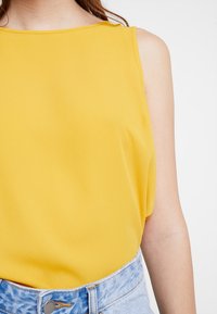 Żółta bezrękawnikowa bluzka z lekkiego materiału o gładkiej teksturze, z okrągłym dekoltem, zestawiona z jasnoniebieskimi szortami dżinsowymi o wysokim stanie.