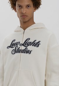 Creme hoodie med frontlynlås, stor kangaroo-lomme og broderet sort tekst "Low Lights Studios." Blød, glat stoftekstur.