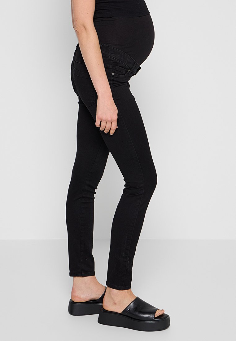 Schwarze Schwangerschaftsjeans aus glattem, dehnbarem Stoff mit gebogener Taillenöffnung; kombiniert mit schwarzen Plateausandalen.