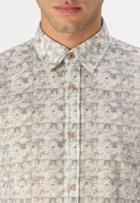 Gepatternde button-up shirt met een lichtbeige en grijs kleurenschema, voorzien van geometrische ontwerpen en bruine knopen. Geweven stofdetail zichtbaar.