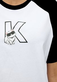 Λευκό και μαύρο μπλουζάκι με μανίκια ράγκλαν, με μεγάλο γράμμα Κ και μια μικρή γάτα που φοράει γυαλιά ηλίου στο στήθος.