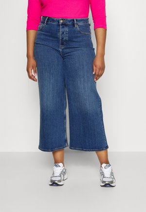 Osoba w szerokich, skróconych niebieskich dżinsach, jaskraworóżowej bluzce z długim rękawem i białych butach do biegania, stojąca na tle jednolitego tła.