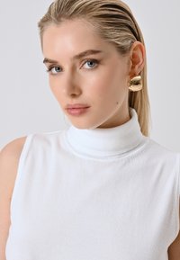 Blonde vrouw met strak naar achteren gekamd haar, gekleed in een witte mouwloze coltrui en grote gouden ringoorbellen, tegen een effen achtergrond.