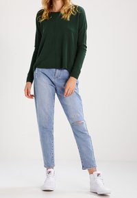 Zielony sweter z długim rękawem o luźnym kroju, zestawiony z jasnoniebieskimi, postarzanymi dżinsami z przetarciem na kolanie oraz białymi trampkami.