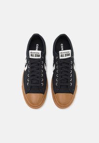 Schwarze Leinensneaker mit einer braunen Gummikappe, weißem Sternlogo und schwarzen Schnürsenkeln. Mit einem Logotag auf der Zunge.