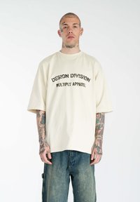 Beige oversized t-shirt med korte ærmer, prydet med sort trykt tekst: "DESIGN DIVISION MULTIPLY APPAREL." Båret med løse denimbukser.