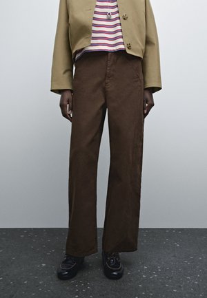 Bruine wijde broek van katoen met zichtbare stikdetails, gecombineerd met een gestreept shirt en een cropped jack. Zwarte schoenen met accenten.