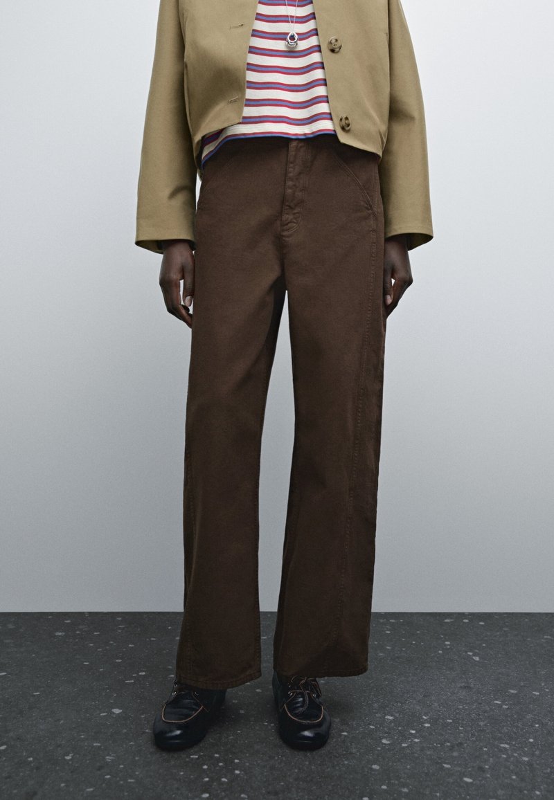 Pantaloni larghi marroni in cotone con dettagli di cucitura visibili, abbinati a una camicia a righe e un giubbotto corto. Scarpe nere con accenti.