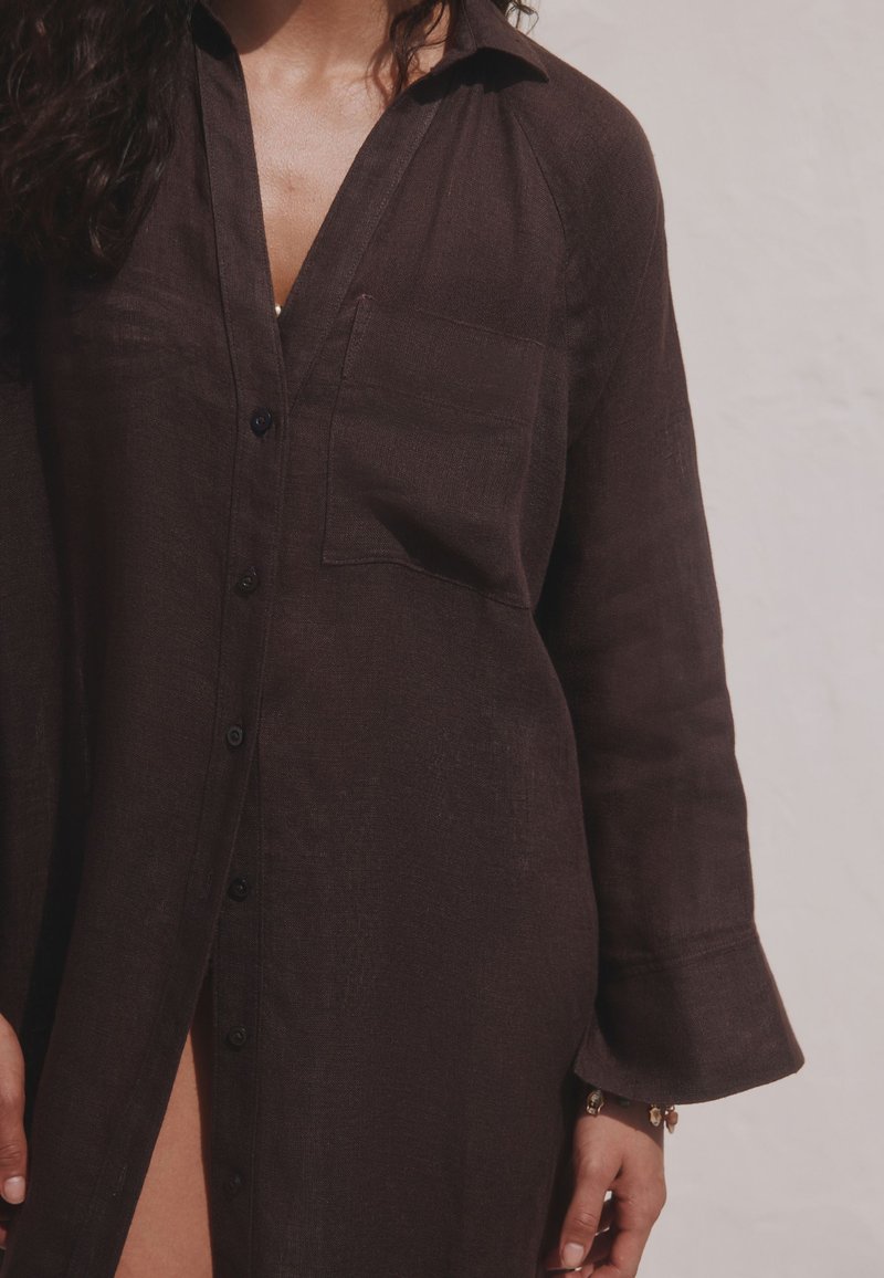 Femme portant une robe-chemise marron foncé à manches longues boutonnée, avec un col et une poche poitrine avant, debout devant un fond clair uni.