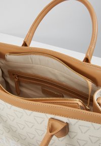 Beige tygväska med bruna läderdetaljer, med ett logomönster, två fack, ett dragkedjefack och dubbla handtag.