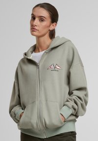 Jasnozielona bluza z kapturem zapinana na zamek, wykonana z mieszanki bawełny, z przednią kieszenią, ribbowymi mankietami i haftowanym wzorem gór w odcieniach czerwieni i niebieskiego.