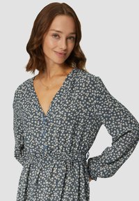 Robe florale bleue à manches longues, décolleté en V, avec des boutons décoratifs et une taille à cordon. Présente un petit motif de fleurs blanches.