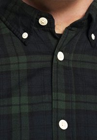 Rutig flanellskjorta i mörkgrön och svart, med vita knappar, krage och en texturerad yta, med överlappande rutmönster.