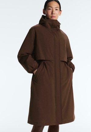 Femme portant un long manteau marron à capuche avec des poignets élastiques, debout les mains dans les poches devant un fond clair uni.