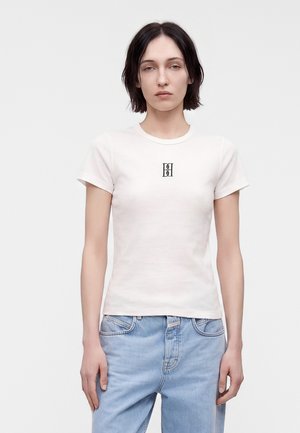 Jeune personne aux cheveux courts et foncés, portant un t-shirt blanc ajusté avec un petit logo noir sur la poitrine et un jean bleu clair.