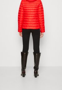 Chaqueta de plumas roja con acolchado horizontal, leggings negros y botas negras hasta la rodilla con suela de tacón y detalles de botón, vistas desde atrás.