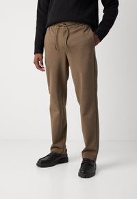 Un pantalon de survêtement marron avec une taille à cordelette, texture lisse, poches latérales et jambes fuselées, associé à des mocassins noirs.