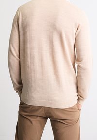 Homme portant un pull beige texturé et un pantalon marron, debout les mains dans les poches, vu de dos sur un fond blanc uni.