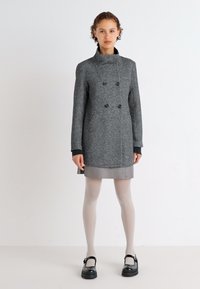 Manteau en mélangé de laine gris avec un col haut, des boutons croisés, et des poches latérales ; associé à une jupe grise et des collants légers.
