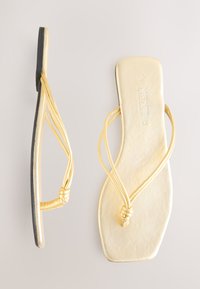 Goldene Flip-Flops mit flacher Sohle, die schlanke Riemen haben, die am Zehensteg verknoten. Glatte metallische Oberfläche und schwarze Gummisohle.