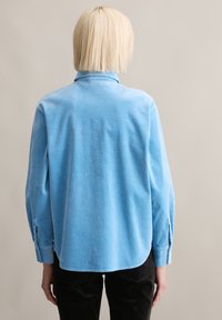 Lichtblauw ribfluwelen overhemd met een omgeplooide kraag, lange mouwen en een licht gebogen zoom, gezien vanaf de achterkant. Textuur van het oppervlak zichtbaar.