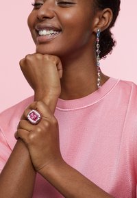 Modelka oblečená v ružovom textúrovanom svetri, ktorá vystavuje veľký ružový drahokamový prsteň a dlhý, farebný krystalový náušník s rôznymi tvarmi a odtieňmi.